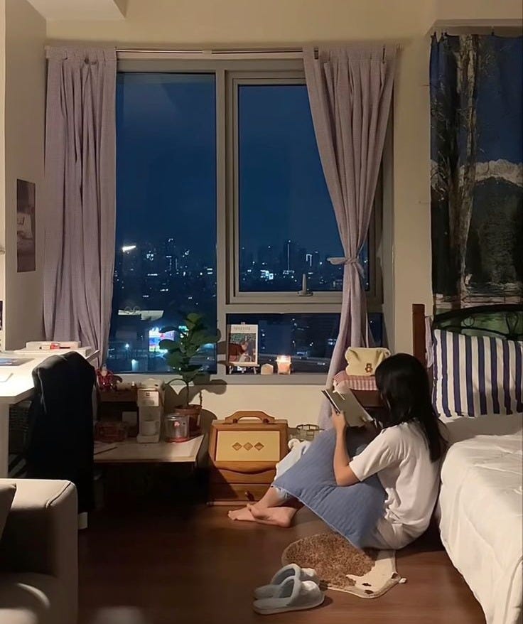 This may contain: a woman sitting on the floor in front of a window looking out at the city
