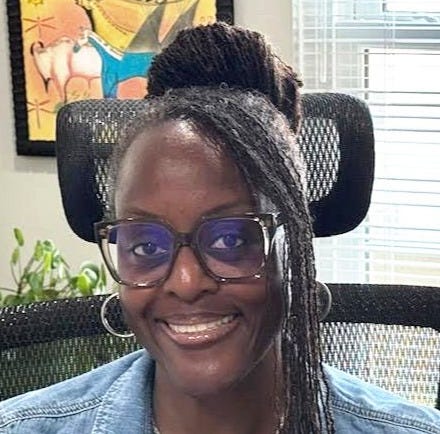 Smiling woman with glasses, wearing a denim jacket, sitting in an office chair, with a framed abstract artwork and window blinds behind her.