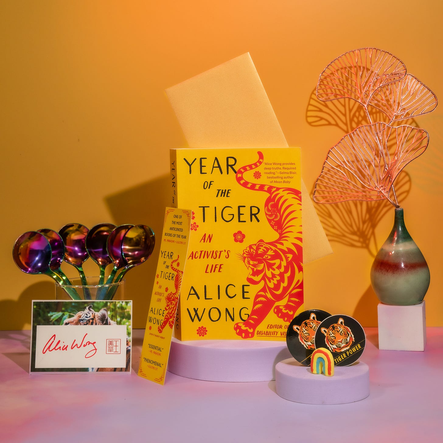 Photo with a tangerine background with a pink border at the bottom with a copy of Year of the Tiger paperback in the center. The left in a glass cup is a bunch of rainbow dipper spoons from @umeshiso_ with a signed tiger bookplate by Alice Wong in front of the cup. A tiger bookmark positioned to the left of the book. On the right is a multicolored pear-shaped vase holding gold ginkgo leaf decorations. In the front is one round tiger sticker and one round tiger button with a small rainbow decor standing upright. Photo credit: @ziru.mo (on IG) Photo with a tangerine background with a pink border at the bottom with a copy of Year of the Tiger paperback in the center. The left in a glass cup is a bunch of rainbow dipper spoons from @umeshiso_ with a signed tiger bookplate by Alice Wong in front of the cup. A tiger bookmark positioned to the left of the book. On the right is a multicolored pear-shaped vase holding gold ginkgo leaf decorations. In the front is one round tiger sticker and one round tiger button with a small rainbow decor standing upright. Photo credit: @ziru.mo (on IG)