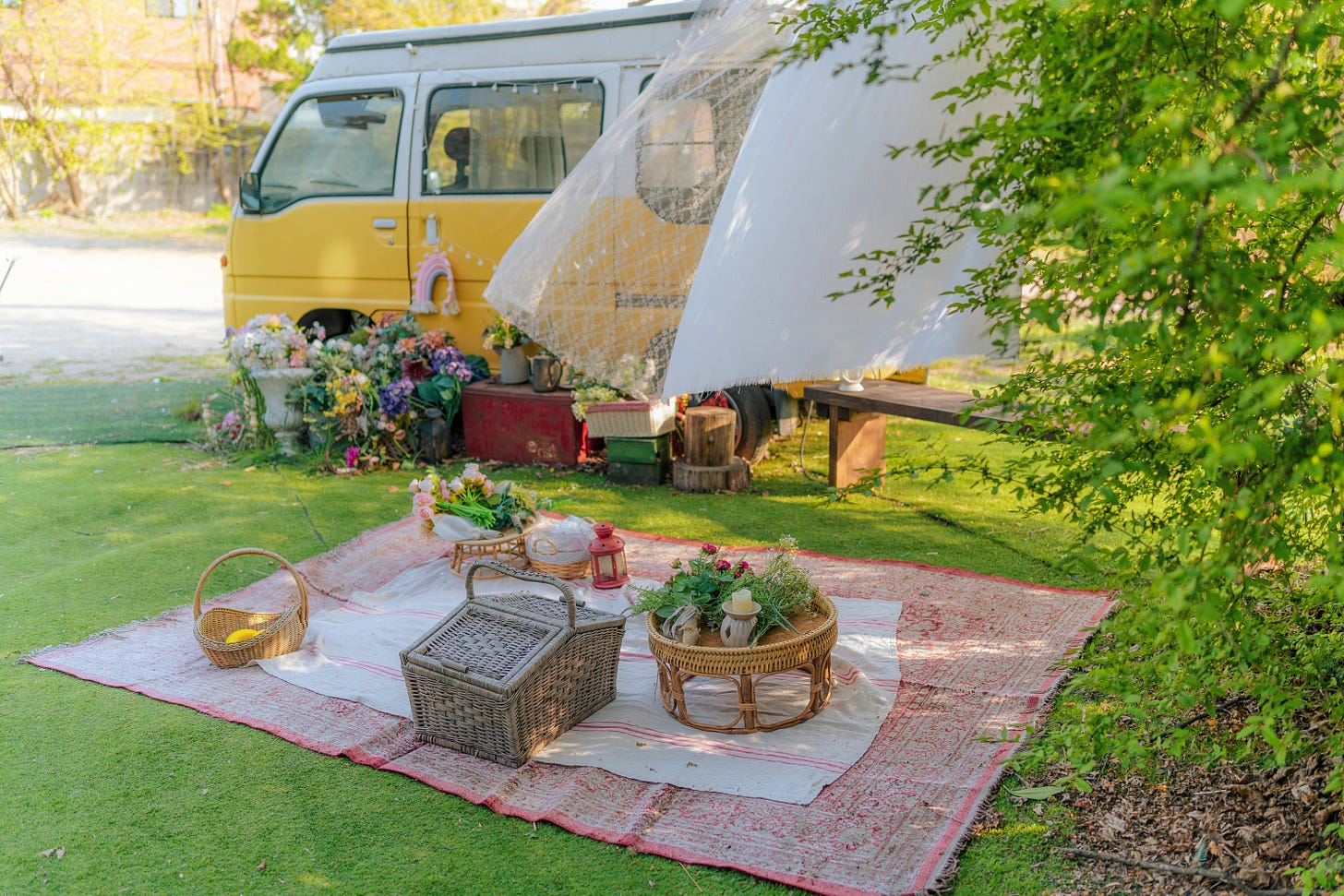Color photo of a picnic laid out on green grass. Behind it is a row of flowers in pots next to a yellow camper van.