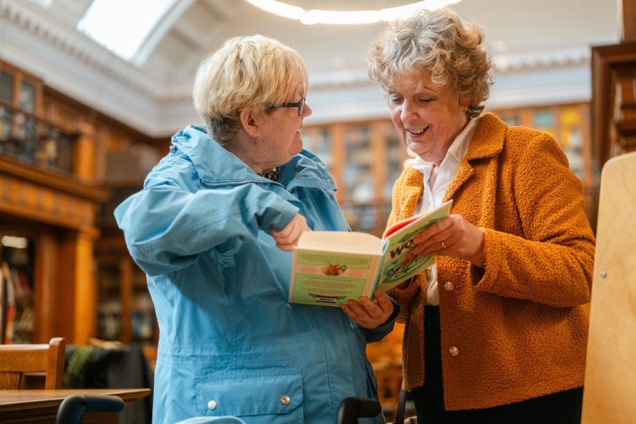 Two older white women, one showing the other something in a book, both smiling.