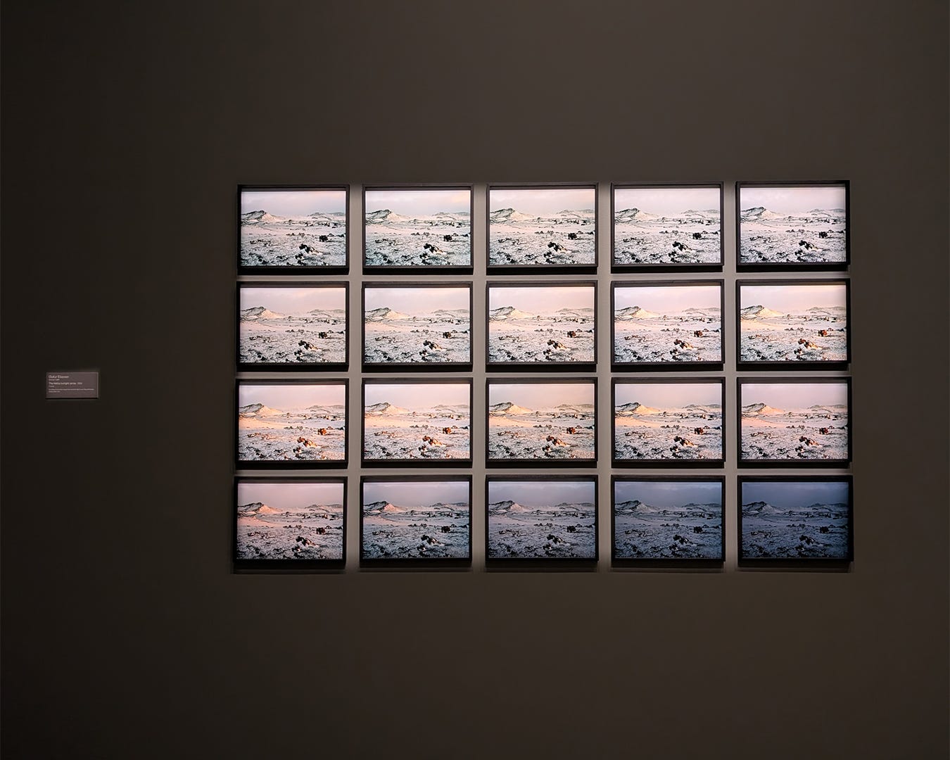 Grid of twenty framed photographs in four rows of five, each showing same Icelandic volcanic landscape under progressively changing light conditions from cool blue-pink dawn tones to warm peachy-orange sunset hues on dark gray gallery wall