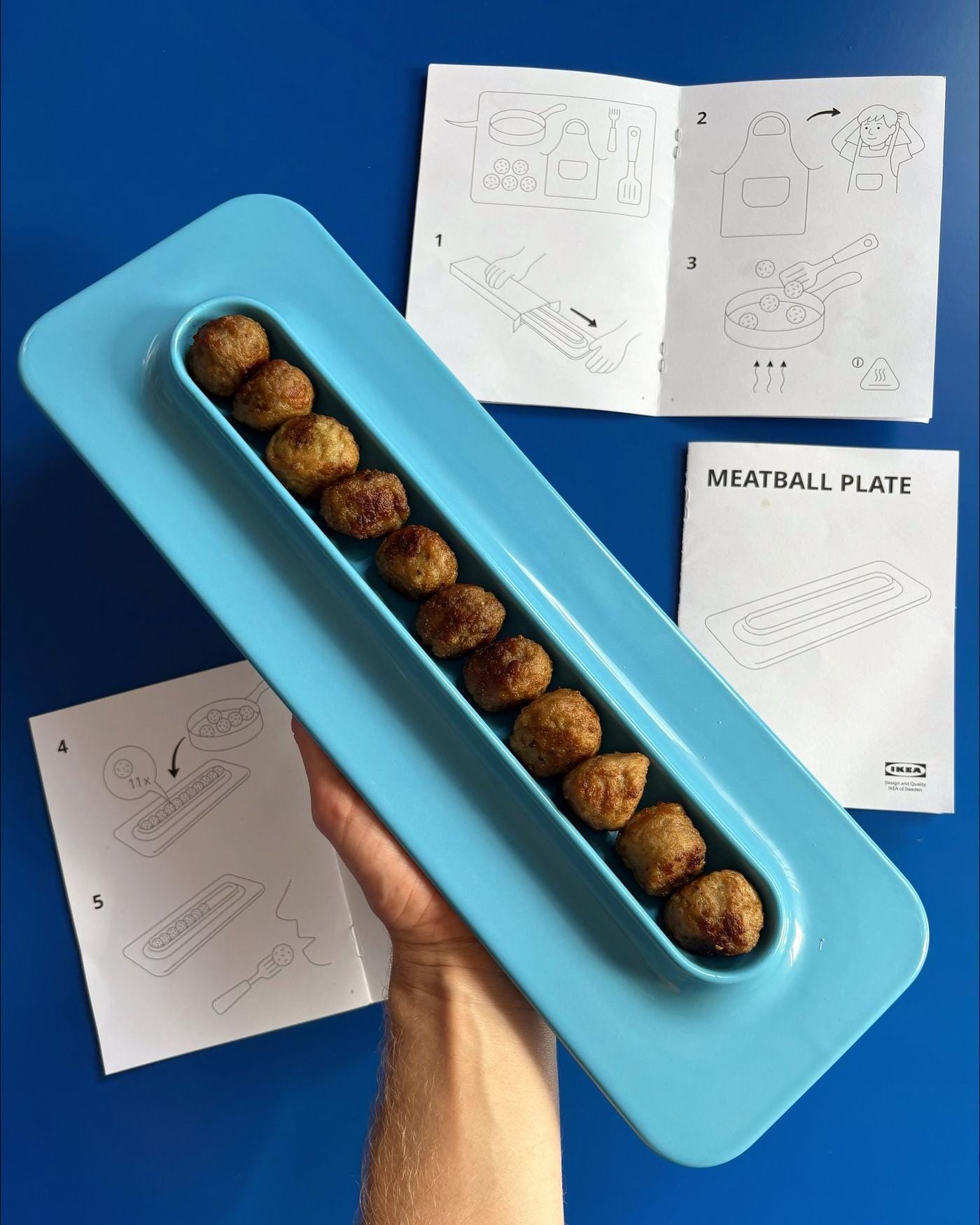 An IKEA instruction manual on a blue surface beside an oblong blue plate held by a hand, with freshly cooked meatballs in a row.