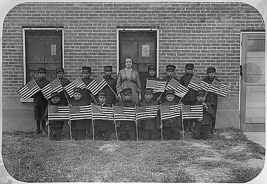 historic black and white photo of 15 uniformed Native American boys, each holding a stiff US flag on a stick, posing in front of a brick school building with their teacher, a white woman who wears a dress. The ground in front of them has sparse patches of grass. Through one window we can see a print of a patriotic painting, possibly of the signing of the Declaration of Independence historic black and white photo of 15 uniformed Native American boys, each holding a stiff US flag on a stick, posing in front of a brick school building with their teacher, a white woman who wears a dress. The ground in front of them has sparse patches of grass. Through one window we can see a print of a patriotic painting, possibly of the signing of the Declaration of Independence
