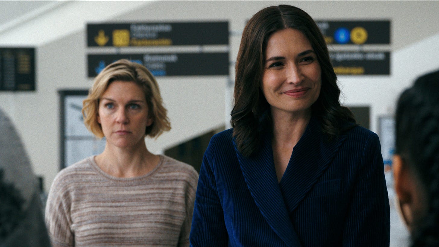 still from pluribus; Carol, blonde, stands looking sad; beside her Zosia, brunette, is smiling and chatting still from pluribus; Carol, blonde, stands looking sad; beside her Zosia, brunette, is smiling and chatting