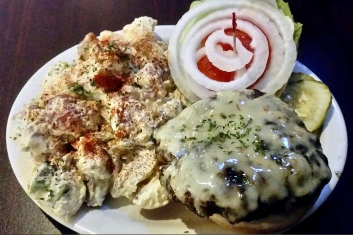 Hand-formed beef patty with melted cheese, onions, and tomato on a bun with a huge side of potato salad.