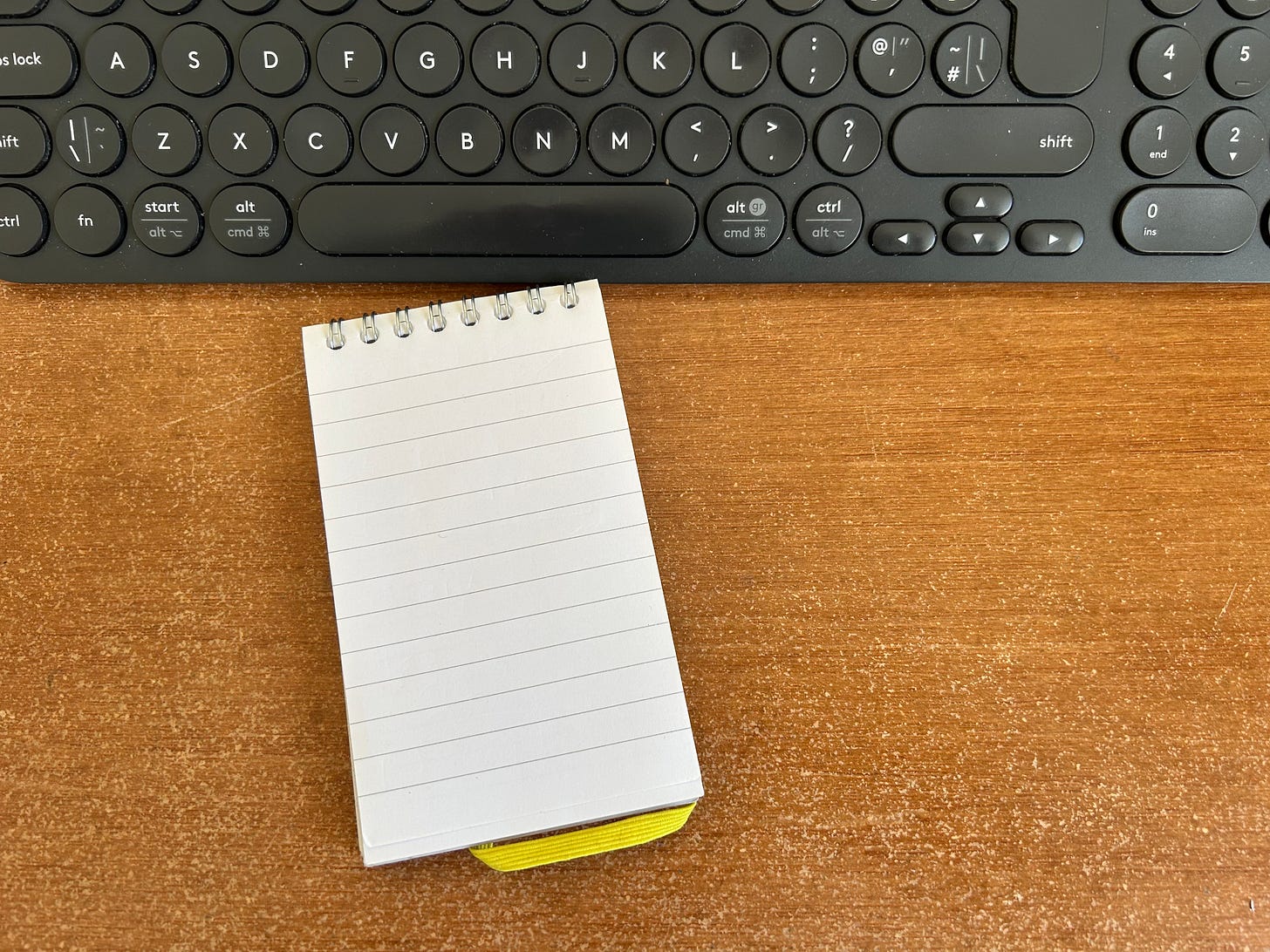 An A7 spiral-bound notebook lying next to a computer keyboard on an office desk.