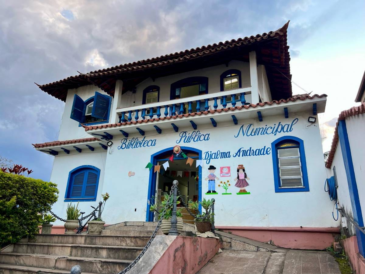 #descrevemospravocê  A imagem mostra a fachada da Biblioteca Pública Municipal Djalma Andrade, localizada em um prédio de dois andares com arquitetura colonial. A construção tem paredes brancas com detalhes em azul, incluindo janelas, portas, beirais e grades da sacada no segundo andar. O telhado é de telhas cerâmicas em estilo colonial, com várias inclinações.  Na parte frontal do prédio, está escrito em letras cursivas azuis: "Biblioteca Pública Municipal Djalma Andrade". Logo abaixo do letreiro, há ilustrações coloridas de duas figuras humanas estilizadas, uma representando um menino com chapéu e outra uma menina de vestido, ambas em trajes típicos de festa junina. Ao centro, entre as duas figuras, está desenhado um vaso com flores. Acima da porta de entrada, há uma pequena decoração com bandeirinhas coloridas.  A entrada principal fica no alto de uma escadaria de pedra, com corrimão de correntes presas a pilares baixos. Vasos de plantas decoram a entrada e há arbustos bem cuidados no lado esquerdo. O céu ao fundo está parcialmente nublado, com tons azulados e cinzentos, sugerindo final de tarde ou tempo instável. A cena transmite acolhimento e preservação da memória local.