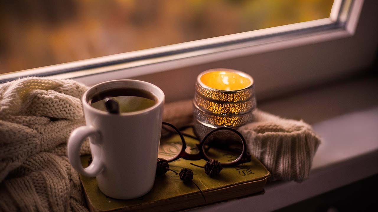 Warm mug and candle resting on a book near a window, with soft autumn light in the background.