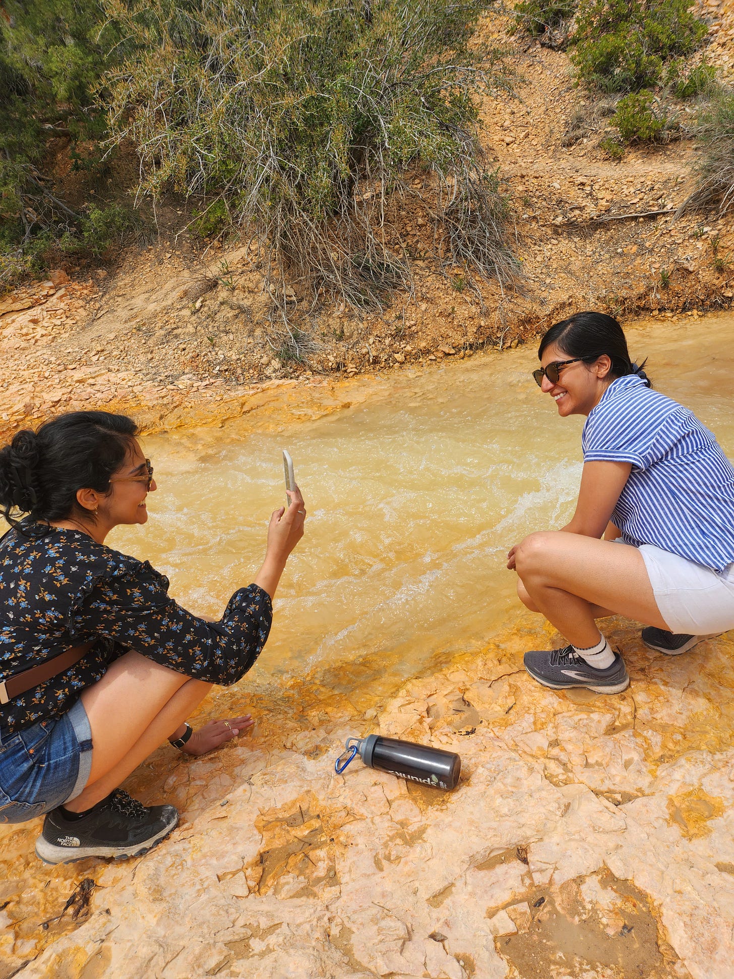 woman in blue blouse and shorts, squatting to take a picture of another woman in white shorts and striped top.