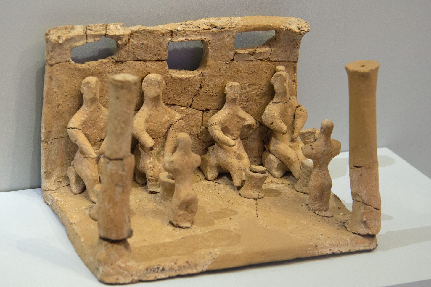 Terracotta model of a small roofless room, open at the sides and front, with one wall at the back containing high windows. There is a column at each of the front corners. Along the back wall sit four schematized human figures on small stools. Each figure has a small table in front of them, with an item on top of it that might be a round loaf of bread. In front of two of the tables are two small humanoid figures that appear to be rising out of the floor.