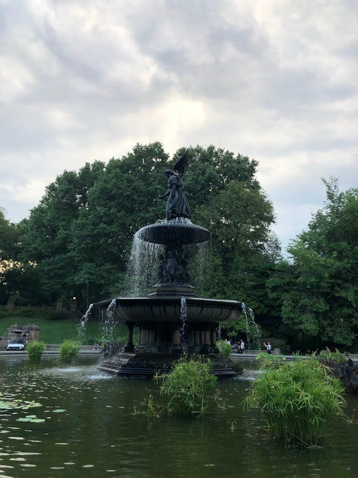 a large, dark green fountain with an angel on top. there are trees behind it, and a cloudy sky.