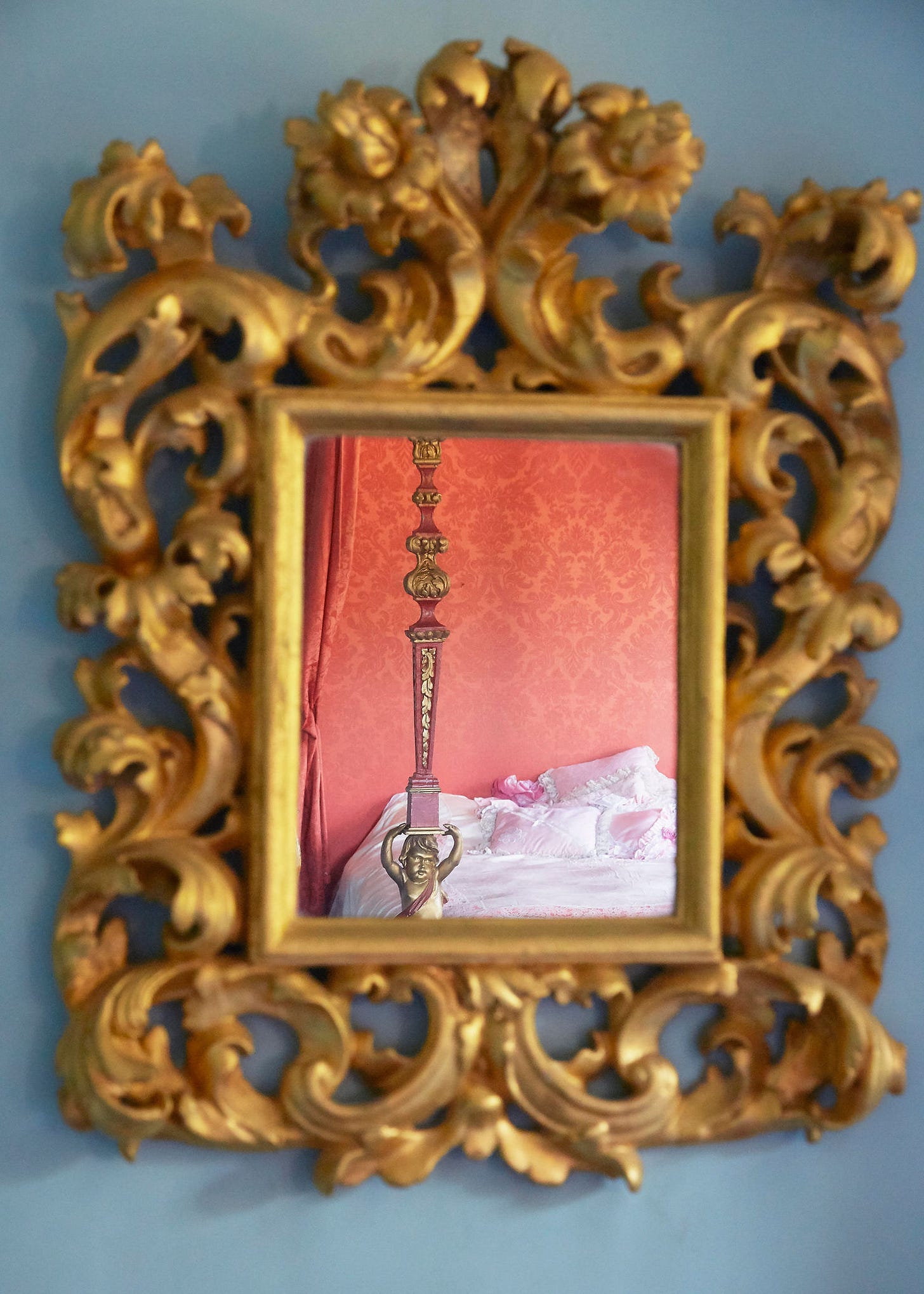 The ornate gold mirror reflects a luxurious bedroom with pink damask wallpaper and a detailed bedpost. It's an elegant, opulent setting. Dame Barbra Cartland's Bedroom Photographed By Claudia Legge
