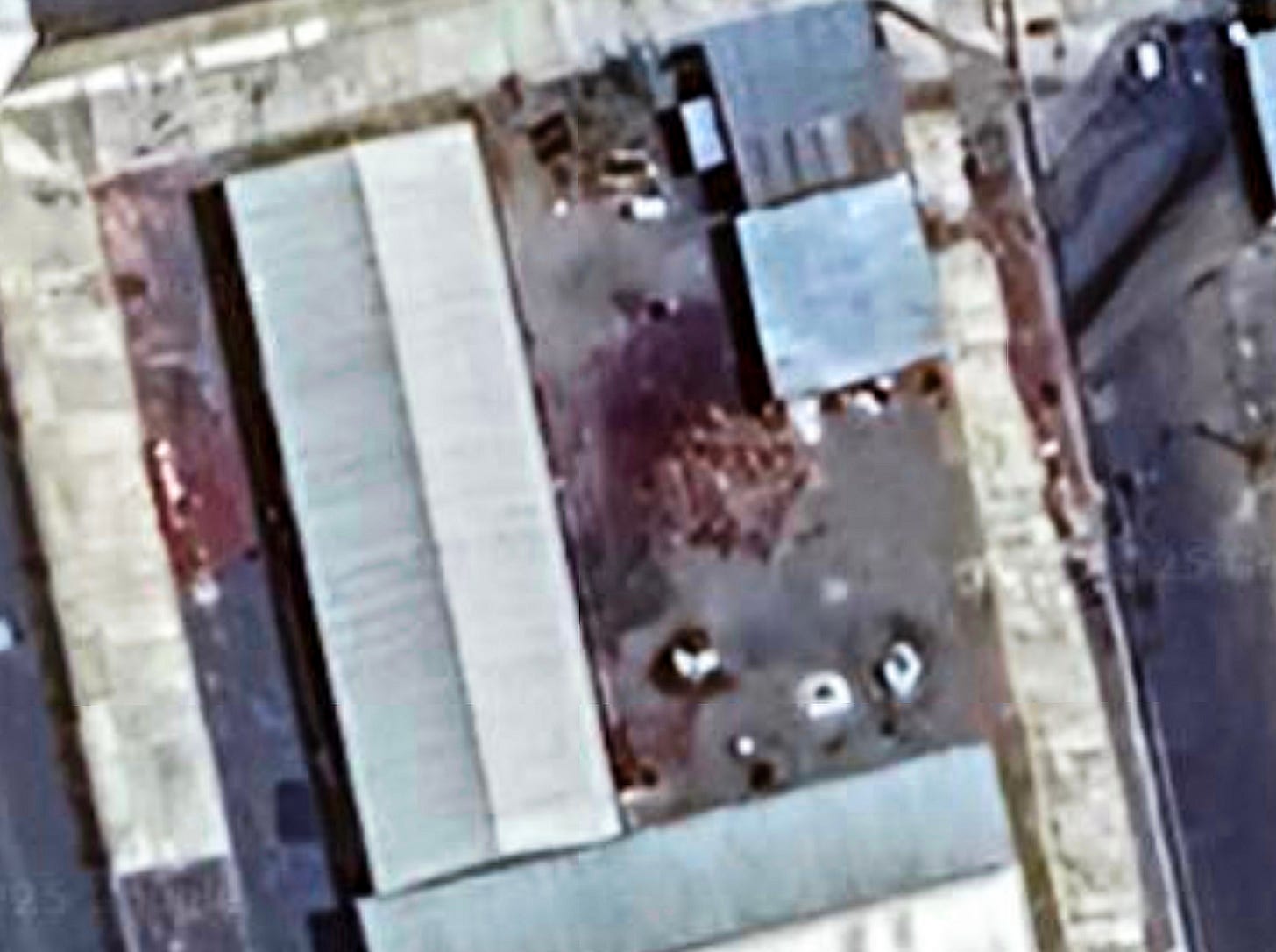 A satellite photo showing the yard of a prison facility with red-stained ground, appearing to be blood. Several small white shapes resembling bodies are visible near the center and lower portion of the yard, surrounded by industrial-looking buildings and paved walkways.