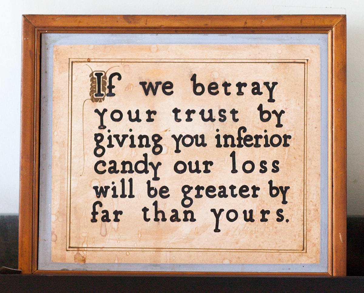 A vintage framed sign with a wooden frame and light blue mat. The aged, cream-colored background has visible water stains. In black typewriter-style font with a decorative illuminated capital "I," the text reads: "If we betray your trust by giving you inferior candy our loss will be greater by far than yours."