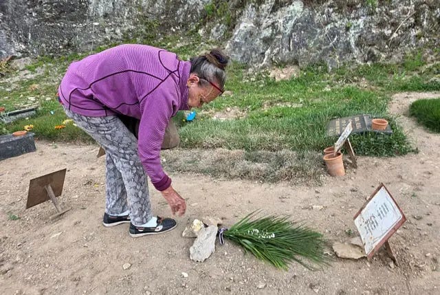 Carmen Nava deposita flores en la tumba de su hijo. Es un suelo de tierra donde la única identificación es una placa metálica con lo que parece un papel encima donde pone el nombre de su hijo. Ella está agachada, tras depositar un ramo que ha dejado pillado con varias piedras. Es una mujer visiblemente mayor, encorvada. Carmen Nava deposita flores en la tumba de su hijo. Es un suelo de tierra donde la única identificación es una placa metálica con lo que parece un papel encima donde pone el nombre de su hijo. Ella está agachada, tras depositar un ramo que ha dejado pillado con varias piedras. Es una mujer visiblemente mayor, encorvada.