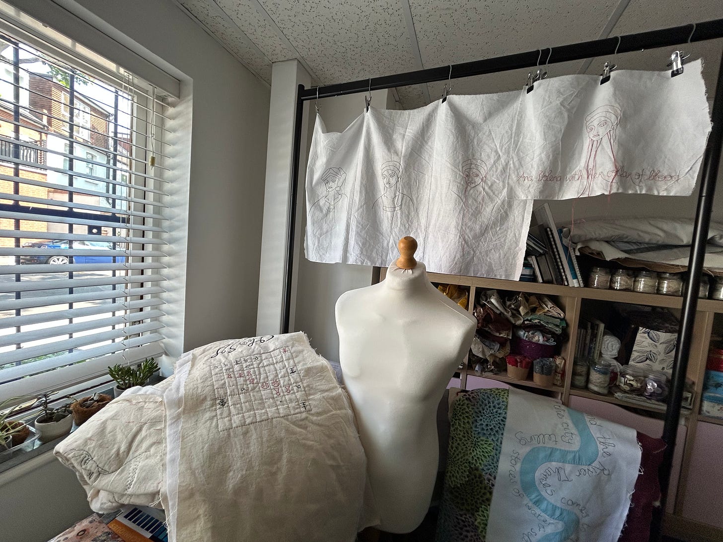 A window with blinds, a tailor’s form, stitched work hanging and resting nearby. Shelving units in the background. 