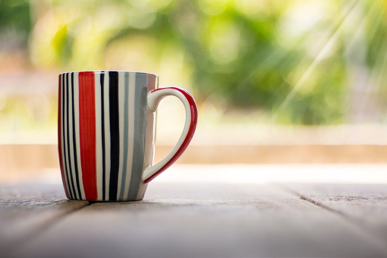 A vertically-striped coffee cup, on a wooden deck, glimmers of sunlight behind it A vertically-striped coffee cup, on a wooden deck, glimmers of sunlight behind it