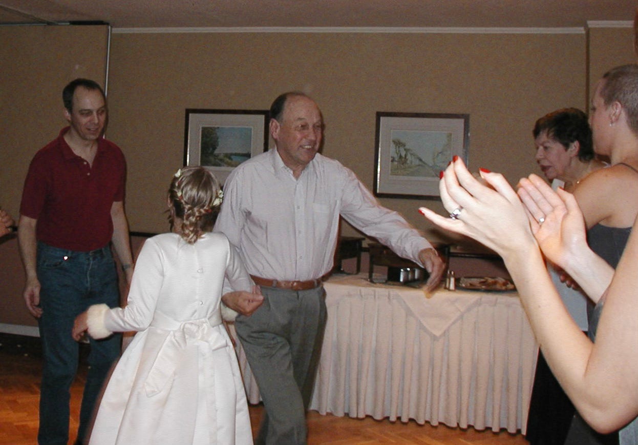 A joyful moment at a wedding in 2001: a smiling older man (the author’s father) dances the eight-some reel with a young girl in a white dress, his step-granddaughter. The author, younger, stands on the left in a maroon polo shirt and jeans, while the author’s mother, a dark-haired woman, watches warmly from the right. Several guests clap along, adding to the celebratory atmosphere in a warmly lit room.