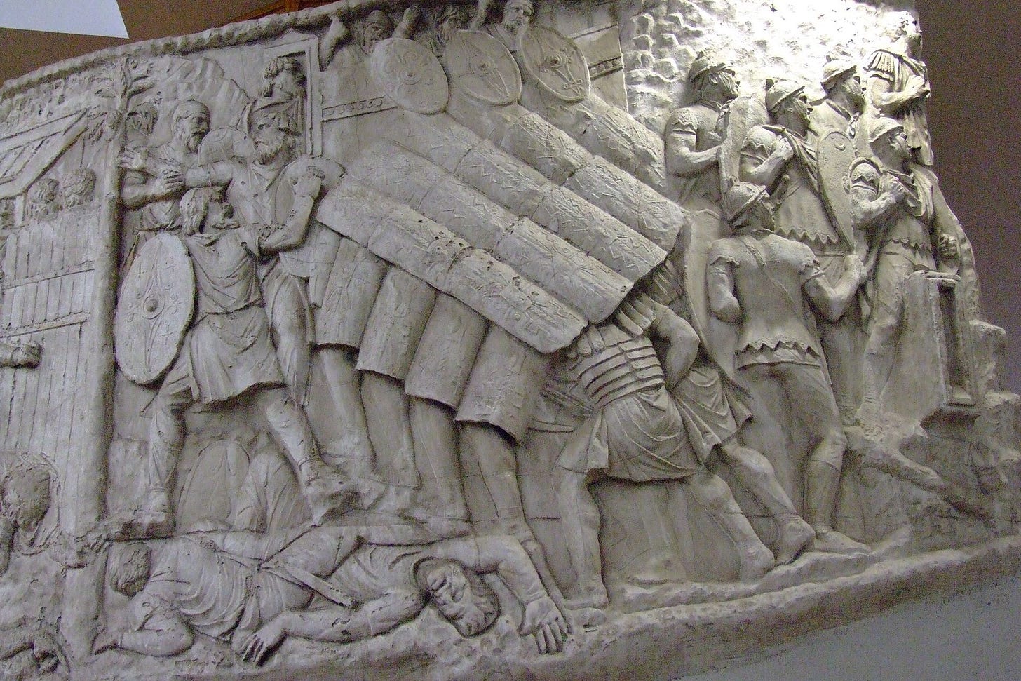 A close-up view of a historical stone bas-relief detail from Trajan's Column, showing a tightly packed unit of Roman legionaries executing a testudo shield wall formation. The soldiers are positioned close together, interlocking their large rectangular shields both vertically to form a front wall and horizontally overhead like overlapping roof tiles to protect themselves against an assault on a Dacian stronghold.