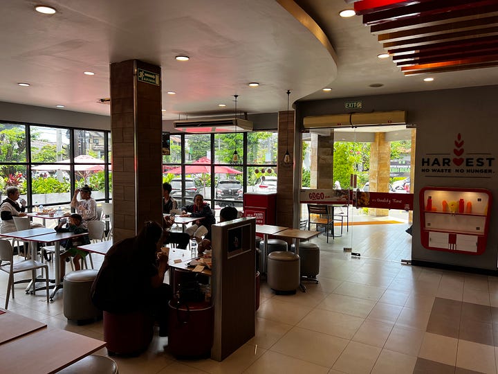  The interior of a KFC, customers at fixed stools, large letters on the wall reading HARVEST and below it NO WASTE NO HUNGER. KFC menu boards advertising rice meal sets, a Kebuli Series, and lychee tea above the service counter. 