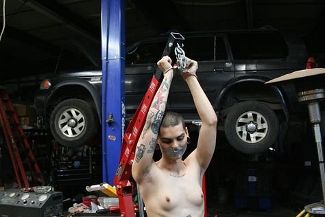A collage of photos of Rae in various forms of bondage: ranging from chained to a car hoist, to rope and rope suspension as well as cuffs and leather bound to a bed, or their ankles. Rae has many tattoos on their arms, legs and stomach. They have a shaved head and brown eyes. They have a large septum ring, a bridge piercing, and stretched earlobes.