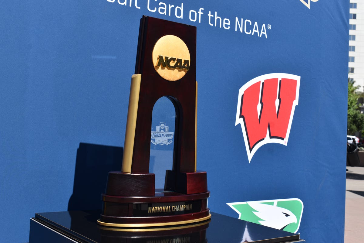 A replica NCAA Tournament trophy sits on a table in front of a backdrop with logos of participating teams including the Wisconsin Badgers logo