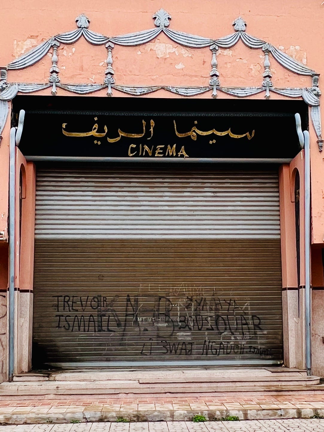 A closed cinema in Marrakech with a metal shutter covered in graffiti, Arabic signage above reading “Cinema, Alrif” and a faded pink facade showing signs of age and disuse. A closed cinema in Marrakech with a metal shutter covered in graffiti, Arabic signage above reading “Cinema, Alrif” and a faded pink facade showing signs of age and disuse.