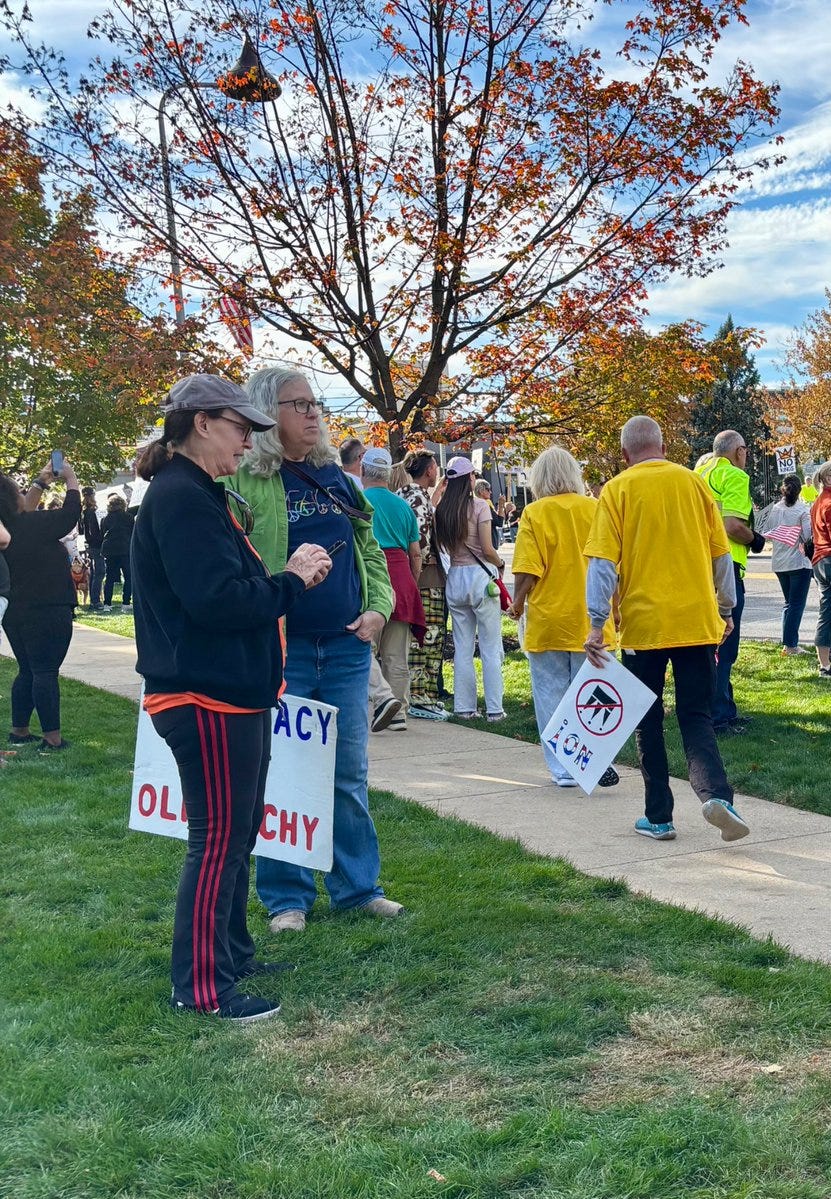 Dr. Rachel Levine stands in a group of protesters at the NoKings event in Hershey PA. Participants hold signs including one with OLDPHACY text and another with a crossed-out symbol. Autumn trees with orange leaves frame the scene, a pumpkin hangs from a branch, people wear casual attire like yellow jackets and red pants, on a grassy path under cloudy sky.