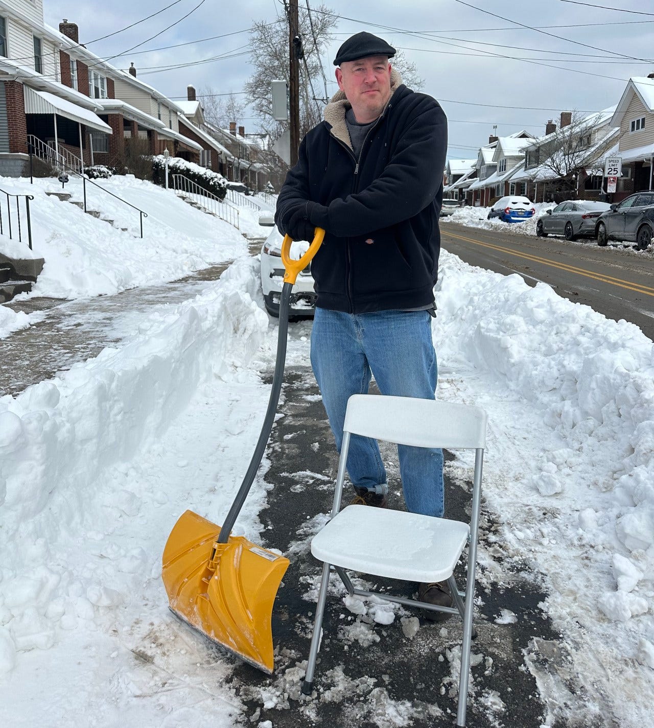 Man standing on a snowy street holding a shovel, next to a white folding chair. Man standing on a snowy street holding a shovel, next to a white folding chair.