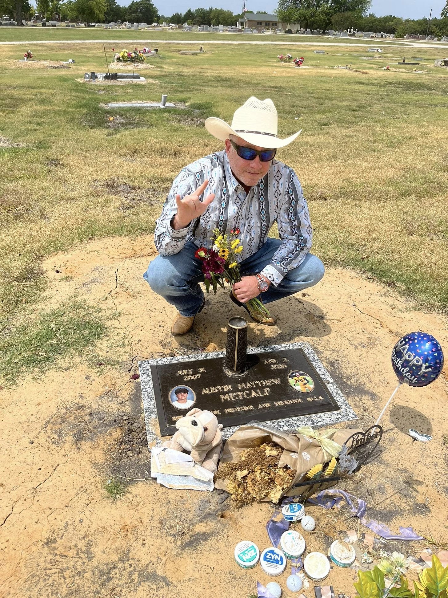 Man in cowboy hat kneeling by a grave marker, holding flowers and celebrating a birthday with mementos and decorations at a cemetery.