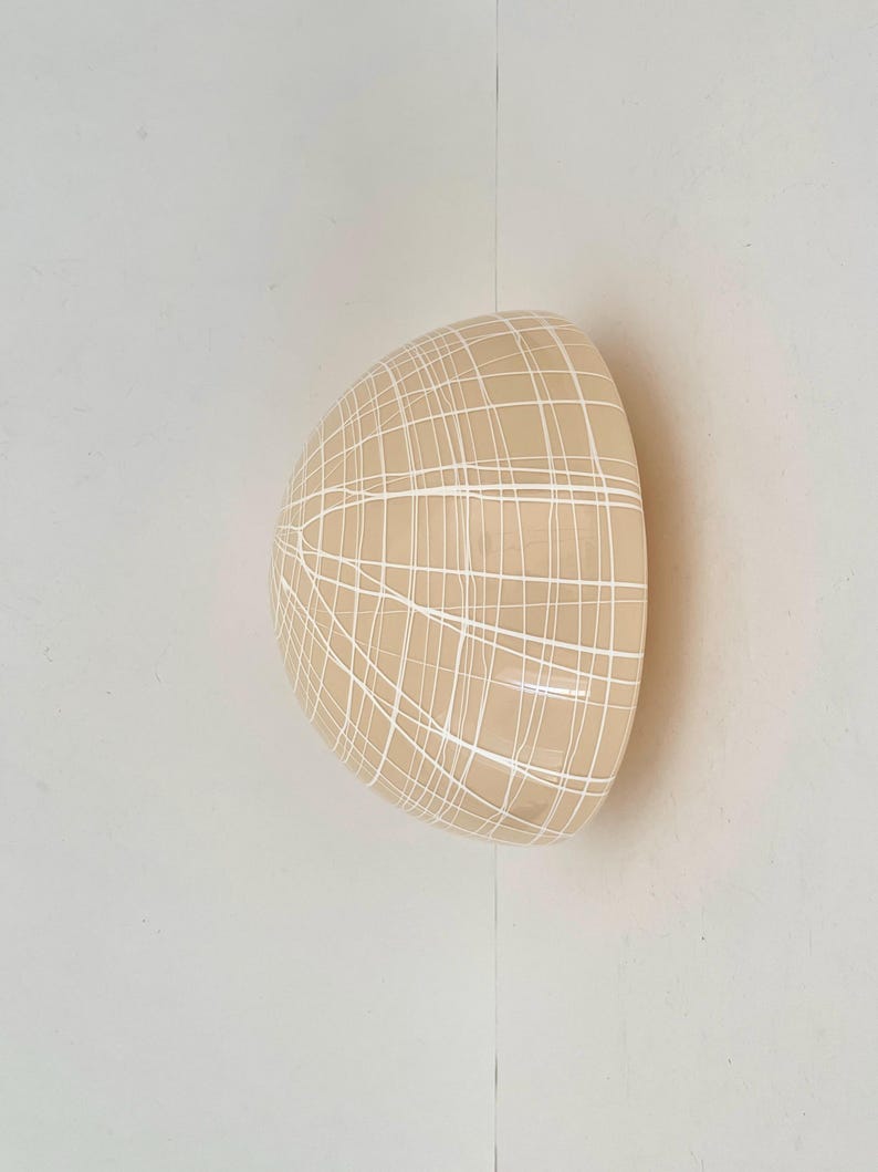 May include: A beige, half-sphere ceramic wall sconce with a white grid pattern. The sconce is mounted on a white wall, creating a minimalist aesthetic. The design is simple and modern.