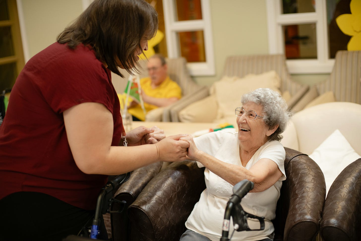 A woman wearing red is holding the hands of an older woman in a white shirt and sitting in a brown leather chair.