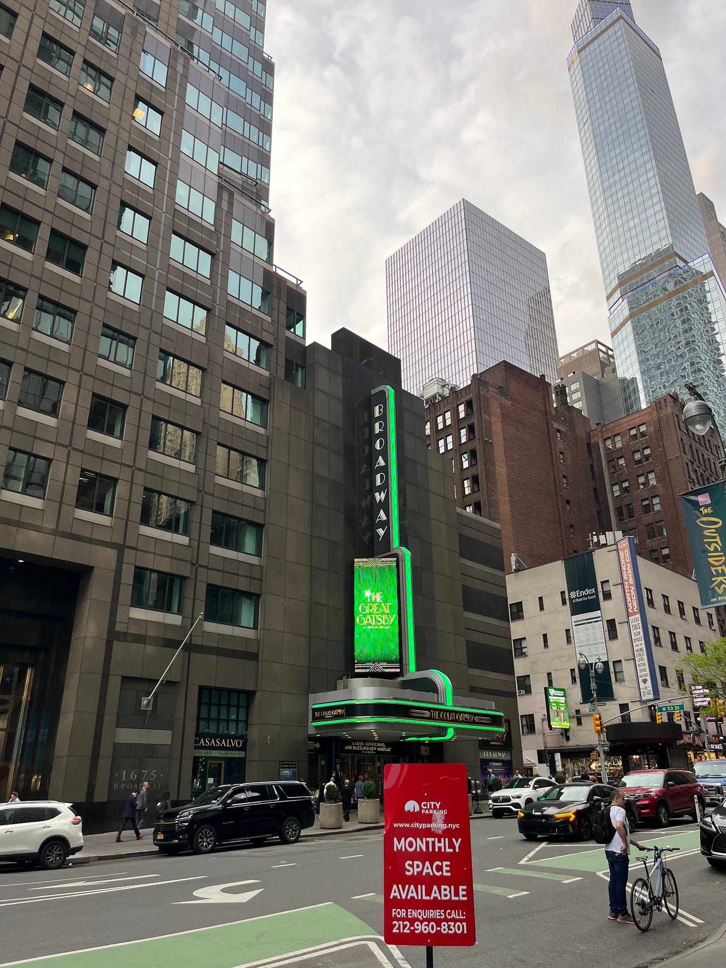 “Broadway” sign outlined in neon green with matching advertisement for “The Great Gatsby,” surrounded by tall buildings and some lightly cloudy sky. A red sign in front reads, “City Parking, Monthly Space Available.” A man in a white t-shirt and backpack, holding his bicycle in his right hand, stands next to the sign, looking into the street.