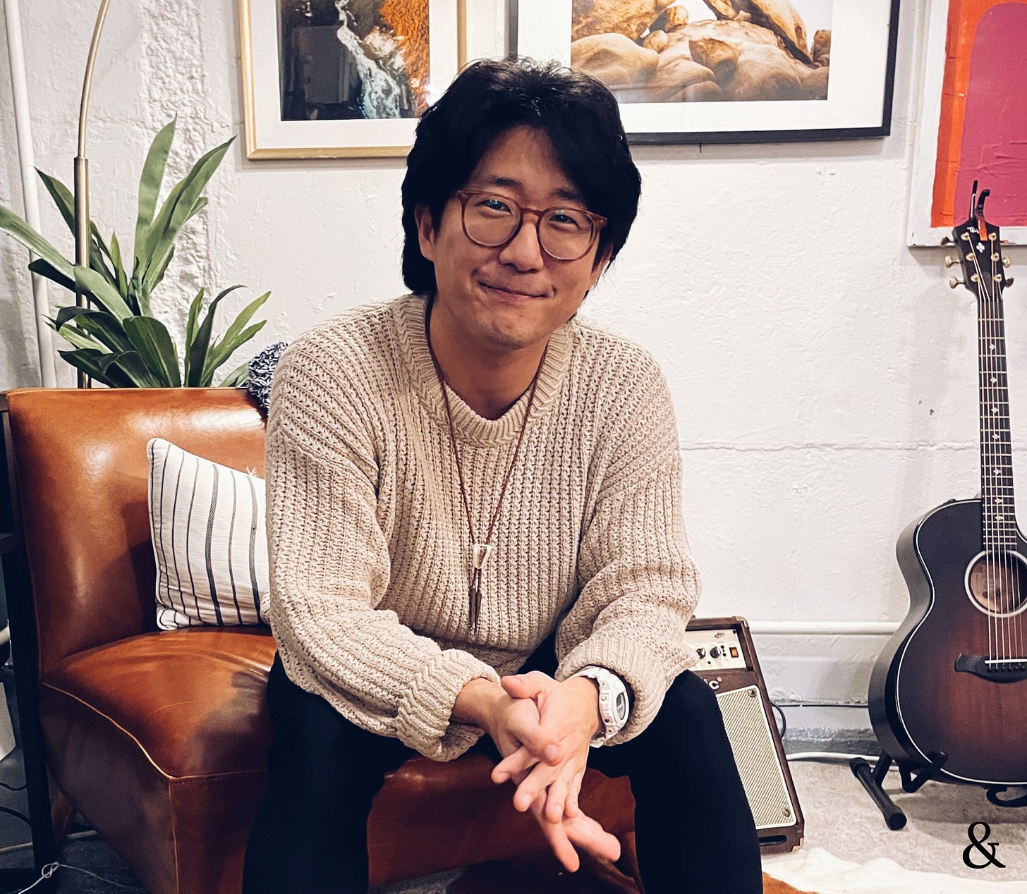 Sam posing with his hands clasped and smiling, while sitting on a leather chair in front of his framed photography, a plant, and a guitar.