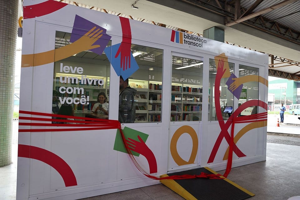 #descrevemospravocê  A imagem mostra uma biblioteca instalada dentro de um contêiner branco, com fachada colorida e janelas amplas. O espaço faz parte do projeto “Biblioteca Transcol”. No canto superior direito da estrutura está o logotipo do projeto, com o nome “biblioteca transcol” escrito em letras pretas ao lado de três faixas verticais nas cores azul, vermelho e laranja. Na janela da esquerda, o texto “leve um livro com você!” aparece em letras brancas.  O contêiner está decorado com formas geométricas e linhas curvas nas cores vermelha, amarela, azul, verde e roxa, que simulam mãos estendidas segurando livros. Uma fita vermelha atravessa a entrada, indicando que o local está prestes a ser inaugurado. Na parte interna, é possível ver estantes organizadas com diversos livros e algumas pessoas no interior da biblioteca.  O espaço está localizado sob uma cobertura metálica, possivelmente em uma estação de ônibus ou terminal urbano. Do lado de fora, há uma rampa de acessibilidade na entrada principal. Ao fundo, vê-se parte da rua e algumas pessoas circulando.