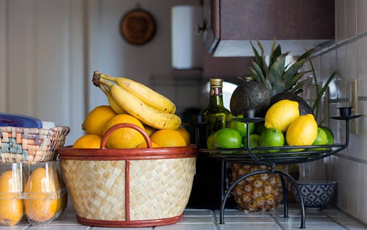 Various fruit sits on a counter: mangos in their plastic container, oranges and bananas in a woven basket, lemons, limes, and avocados in a wire fruit dish/candle holder combo, with a pineapple peaking out from behind.  The counter top and backsplash are white tile.