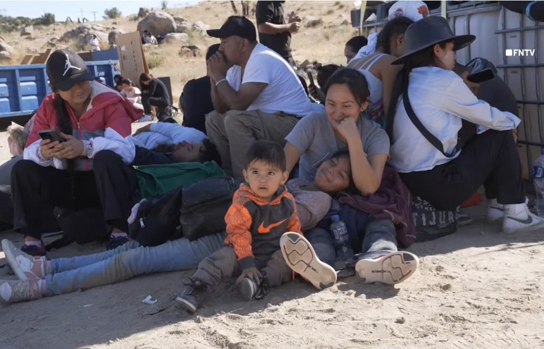 Video screenshot of about a dozen migrants sitting in what shade they can find next to a stack of supplies on pallets in the desert sand. In the foreground sit a young mother, a tween girl lying in the sand with her head on the woman' lap, and a toddler in sneakers, pants, and an orange and black long-sleeved hoodie that's out of place in the June heat. The children's clothing is dusty because they're sitting on the bare dirt. Other people sit nearby, some looking at phones, napping, or staring into space. Behind this group, other clusters of people sit, some in the shade of rocks on a hillside, others in full sun. Video screenshot of about a dozen migrants sitting in what shade they can find next to a stack of supplies on pallets in the desert sand. In the foreground sit a young mother, a tween girl lying in the sand with her head on the woman' lap, and a toddler in sneakers, pants, and an orange and black long-sleeved hoodie that's out of place in the June heat. The children's clothing is dusty because they're sitting on the bare dirt. Other people sit nearby, some looking at phones, napping, or staring into space. Behind this group, other clusters of people sit, some in the shade of rocks on a hillside, others in full sun.
