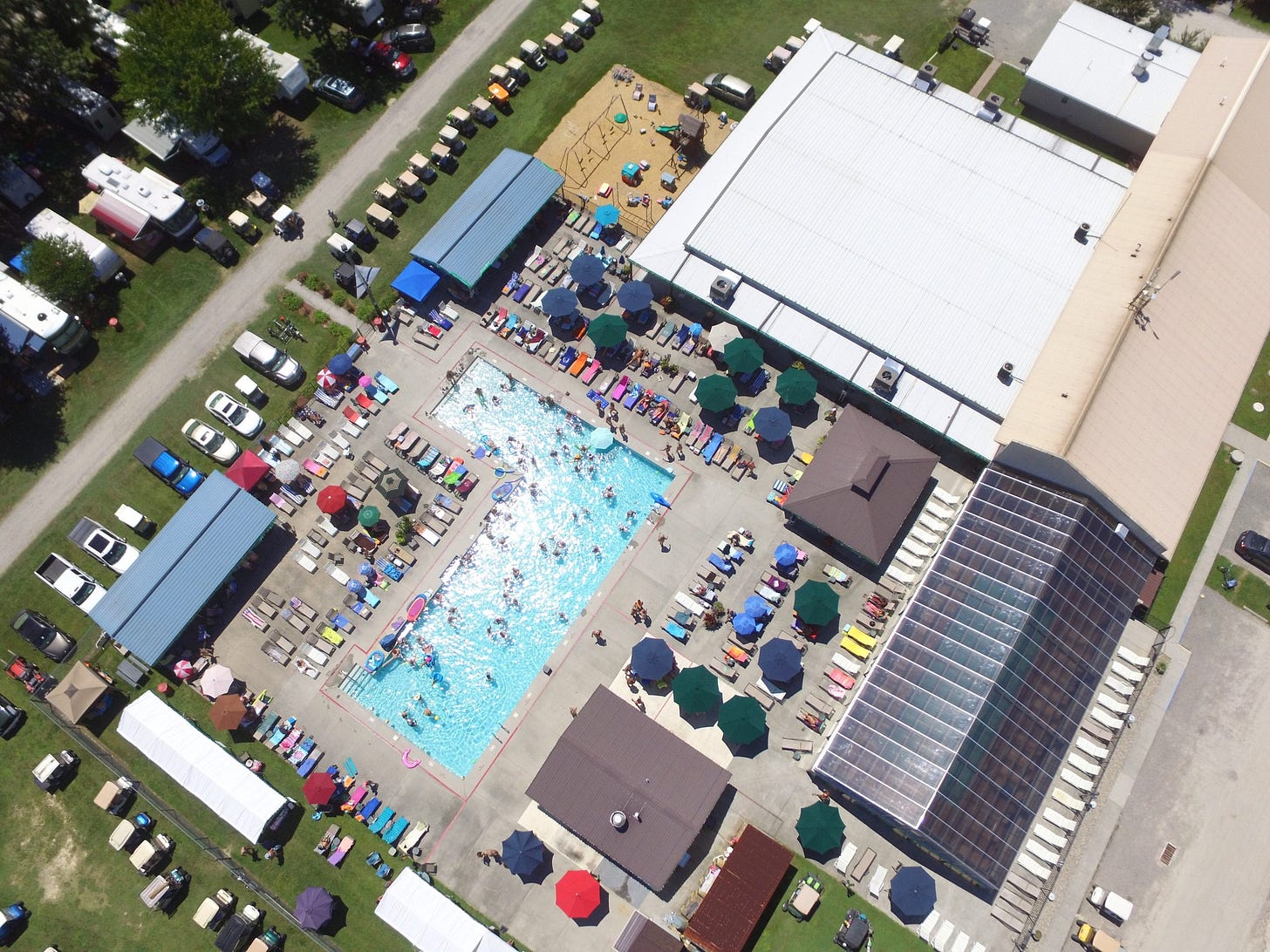 Aerial view of White Tail Resort: a large rectangular outdoor pool ringed by sun loungers and umbrellas, with an enclosed pool building, clubhouse, playground, RV sites, and golf carts visible across the grounds.