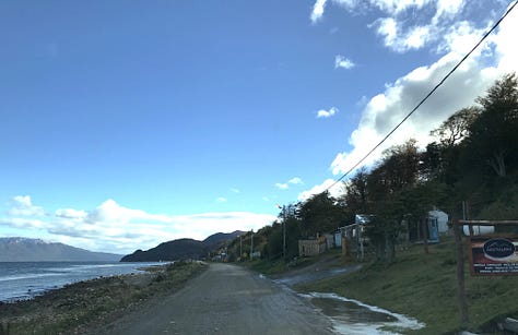A road bordered by buildings, facing the water; a man's hands pointing through the windshield with a machine gun visible through the driver's window; a fishing dock with mountains and a town on the other side of a stretch of water.