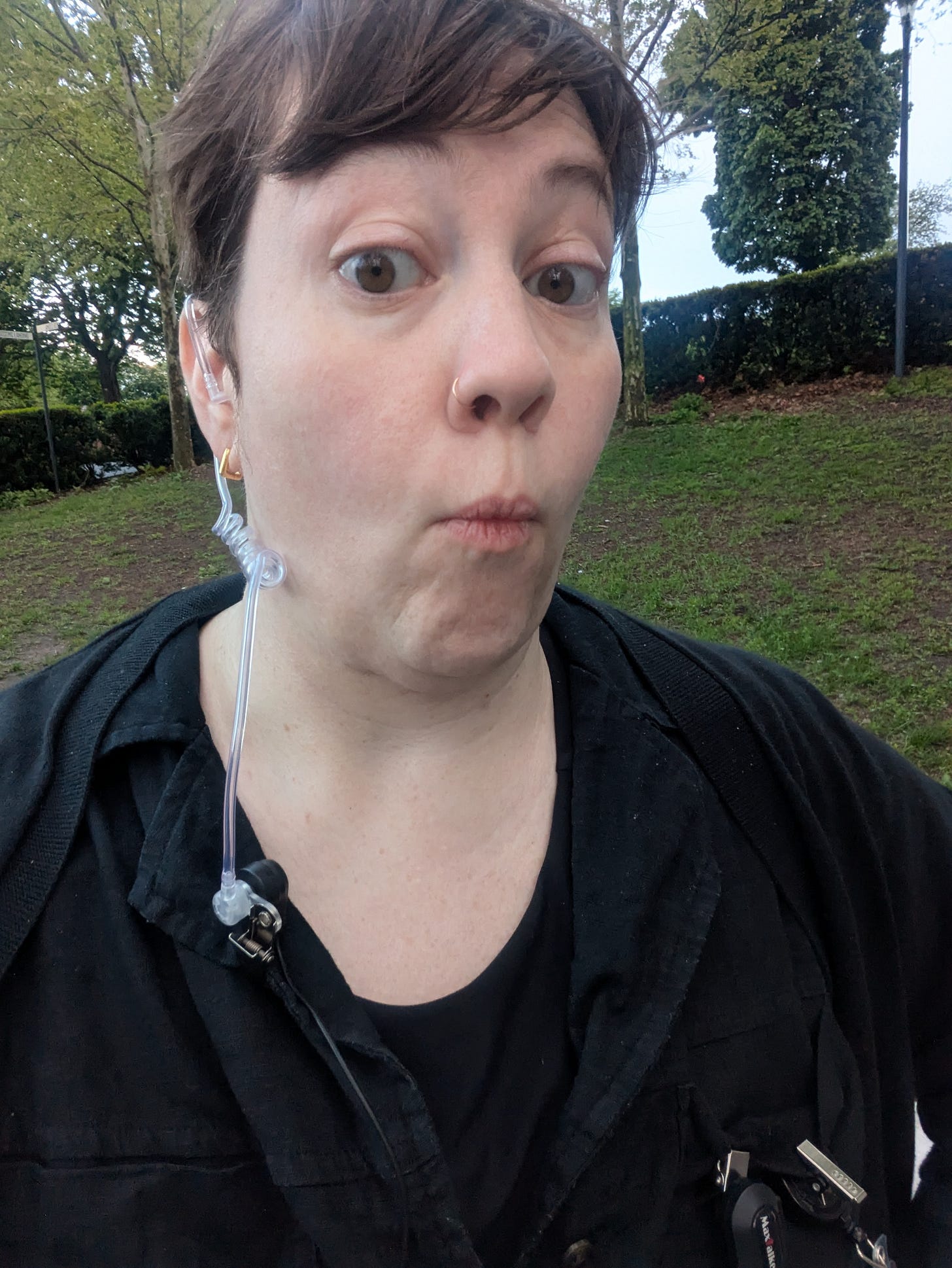 the author looking at the camera while wearing a clear plastic ear piece standing in front of shrubs and greenery