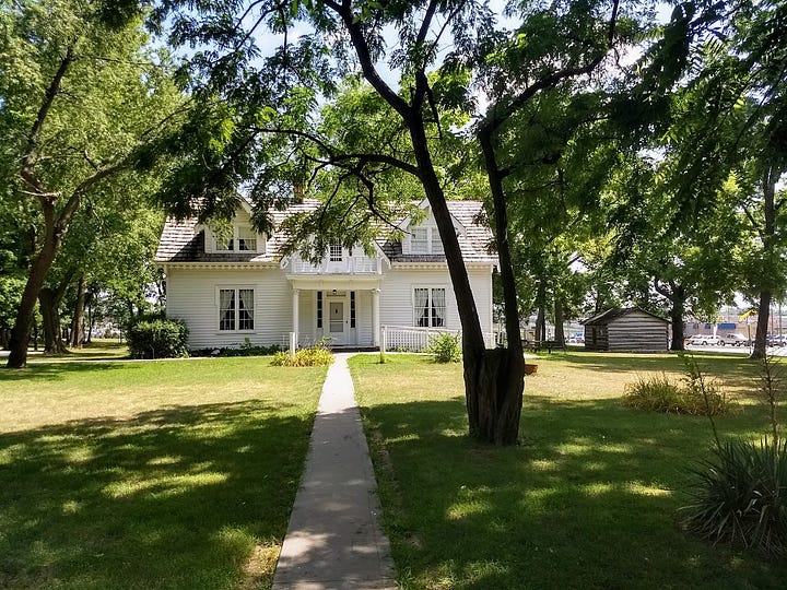 Modest one story house with attic rooms set in large lawn with cars and development glimpsable across the main road plus historic signs