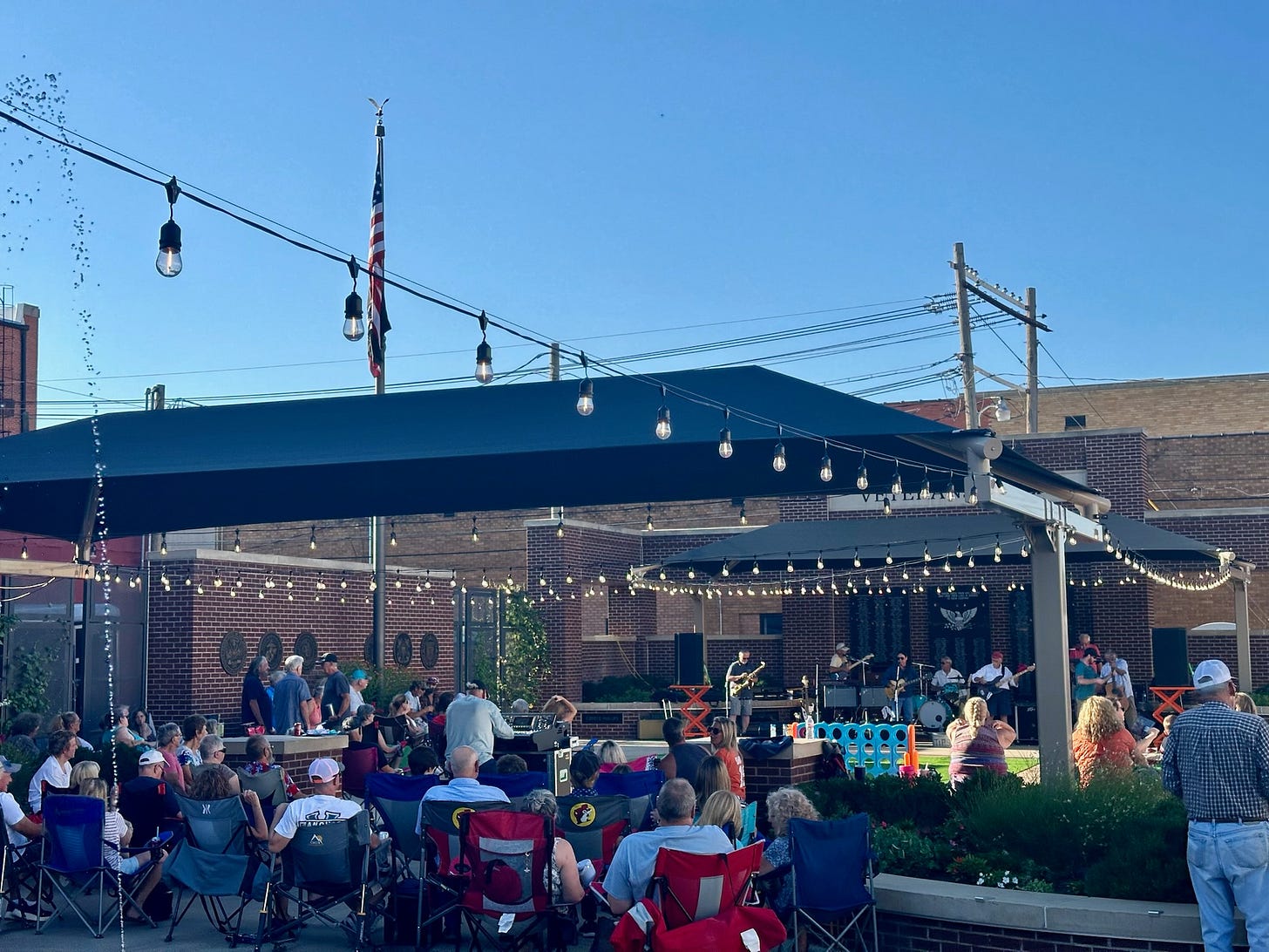 A large group of people are sitting outside under a canopy, enjoying a live music performance.