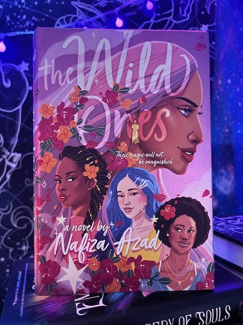 A book propped on a stack of books with glowing purple and blue lights and tapestries in the background. The book is the Wild Ones, a novel by Nafiza Azad with four young women in the cover art, one bigger than the others with her profile to the side, and the three below in a row looking out at the reader. The cover has shades of purple and has decorations of stars and flowers. A book propped on a stack of books with glowing purple and blue lights and tapestries in the background. The book is the Wild Ones, a novel by Nafiza Azad with four young women in the cover art, one bigger than the others with her profile to the side, and the three below in a row looking out at the reader. The cover has shades of purple and has decorations of stars and flowers.