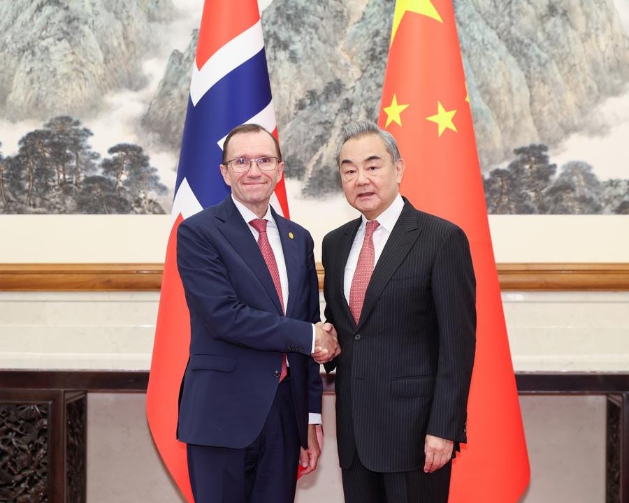 Two men in dark suits and red ties shake hands in front of Norwegian and Chinese flags, with a mountainous landscape painting on the wall behind them in a formal room setting.