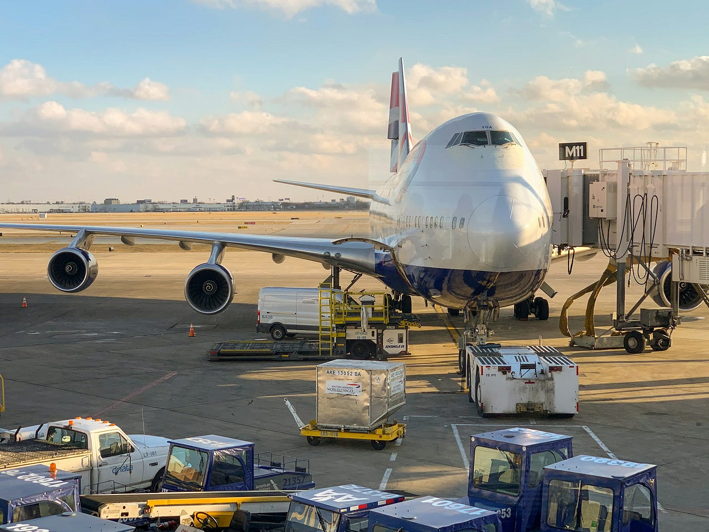 A British Airways Boeing 747 aircraft parked at gate M11 at Chicago O'Hare International Airport Terminal 1. The plane is connected to a jetway with various ground service vehicles and cargo containers surrounding it. The image shows a clear day with blue skies and scattered clouds, with the airport tarmac visible.
