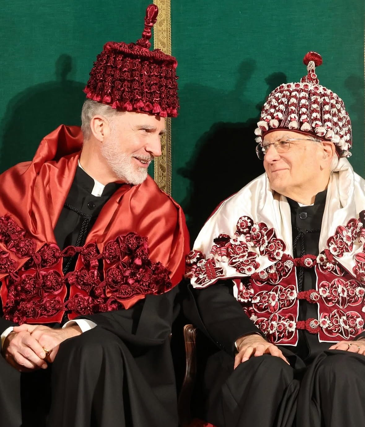 Due uomini in abiti accademici sono seduti uno di fronte all'altro, intenti a conversare. L'uomo a sinistra ha la barba grigia e indossa una toga nera con un vistoso cappuccio accademico rosso, caratterizzato da intricate balze rosse a strati e decorazioni. Indossa anche un cappello rosso abbinato con una texture simile a balze e una punta. L'uomo a destra, che porta gli occhiali, è vestito con una toga nera e un cappuccio accademico bianco e rosso. Il suo cappuccio è adornato con applicazioni floreali bianche e rosse e un bordo decorativo. Indossa un cappello bianco con accenti rossi, caratterizzato da un design arrotondato e testurizzato e un piccolo ornamento rosso sulla parte superiore. Lo sfondo è un tessuto verde scuro, probabilmente una tenda o un rivestimento murale.
