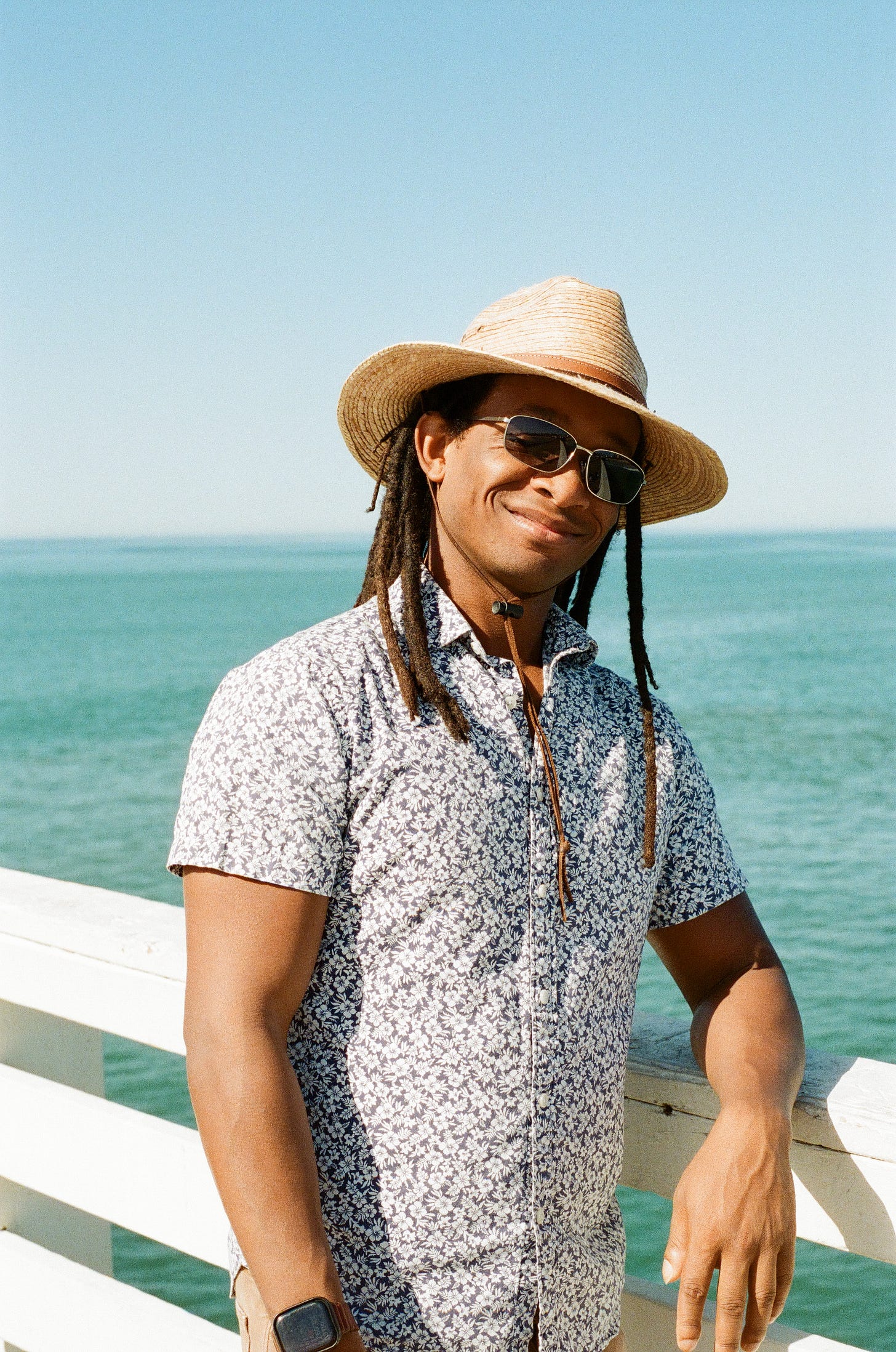 Dapper dressed San Diegan with hat and sunglasses in a stylish short sleeved button down leaning against the pier, with a smile that says he knows how good he looks...but in a nice way