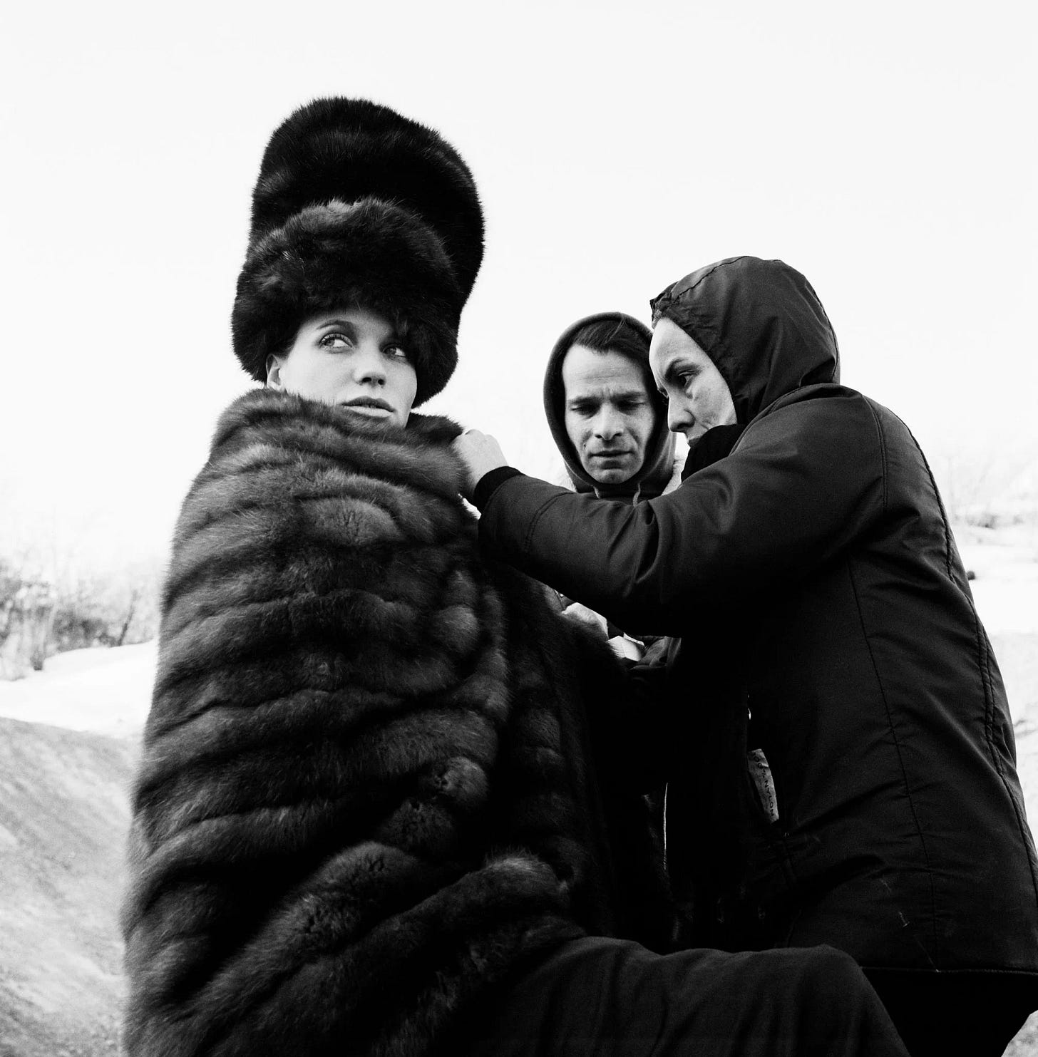 A fur-clad woman is being dressed by another, smaller woman wearing a hooded jacket as a man beside them.