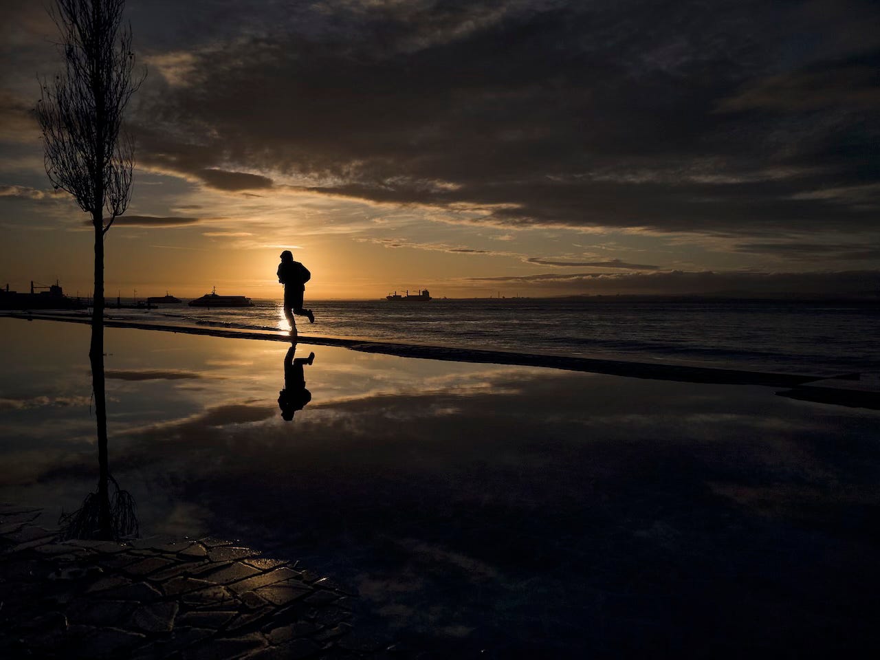 The silhouette of a runner is detached against the sun rising over the river and reflected in a wide puddle on Ribeira das Naus in Lisbon The silhouette of a runner is detached against the sun rising over the river and reflected in a wide puddle on Ribeira das Naus in Lisbon